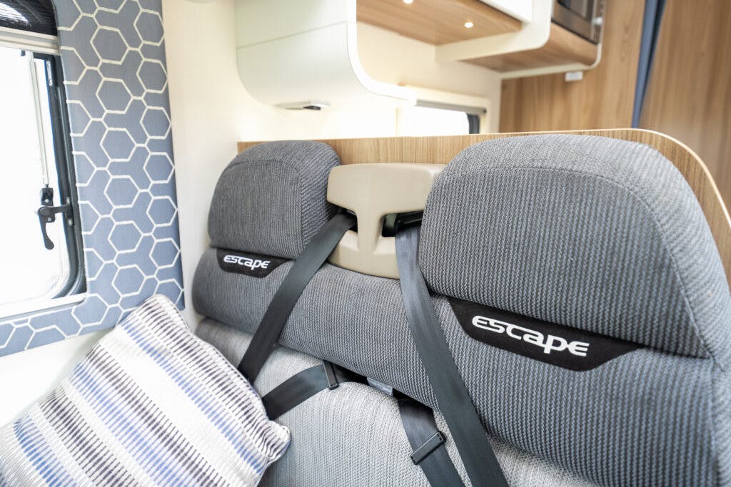 Interior view of a 2017 Swift Escape 664 RV with gray headrests labeled "Escape," gray and white patterned cushions, and hexagon-patterned window curtains on the left. The background features wooden cabinetry and overhead storage.
