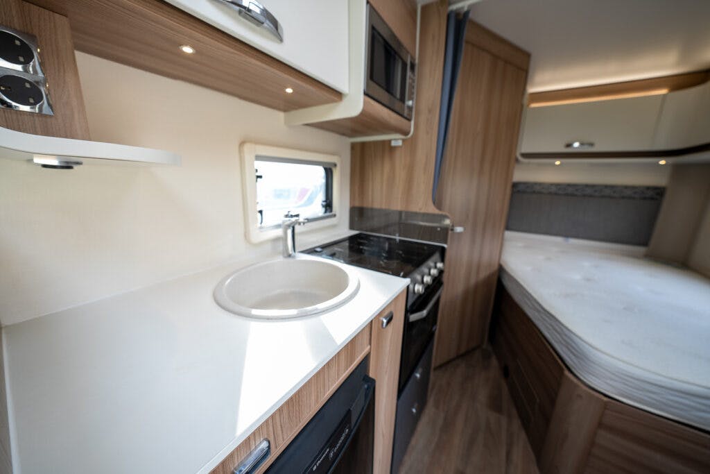 The image shows the interior of a 2017 Swift Escape 664 RV. It features a kitchenette with a sink, stove, microwave, and cabinets. Adjacent to the kitchen area is a small bed with white linens. The design is modern with wood finish and neutral colors.
