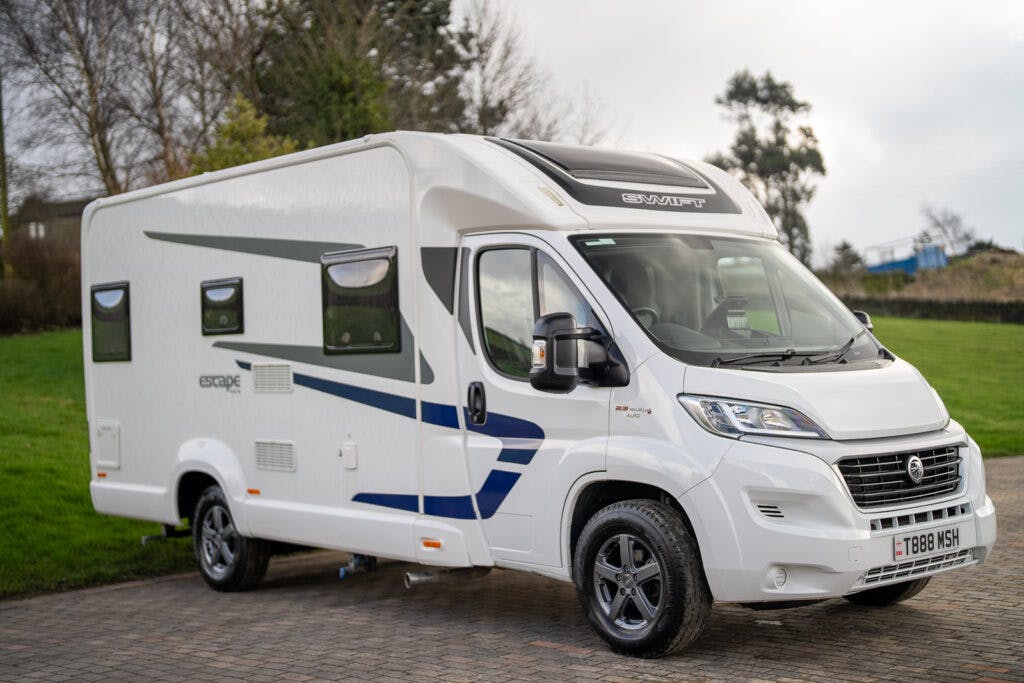 A 2017 Swift Escape 664 motorhome with the brand name "Swift" on the front, parked on a paved area. The motorhome boasts a blue stripe design along the side, tinted windows, and a UK license plate reading "T188 MSH". Trees and greenery provide a lush backdrop.