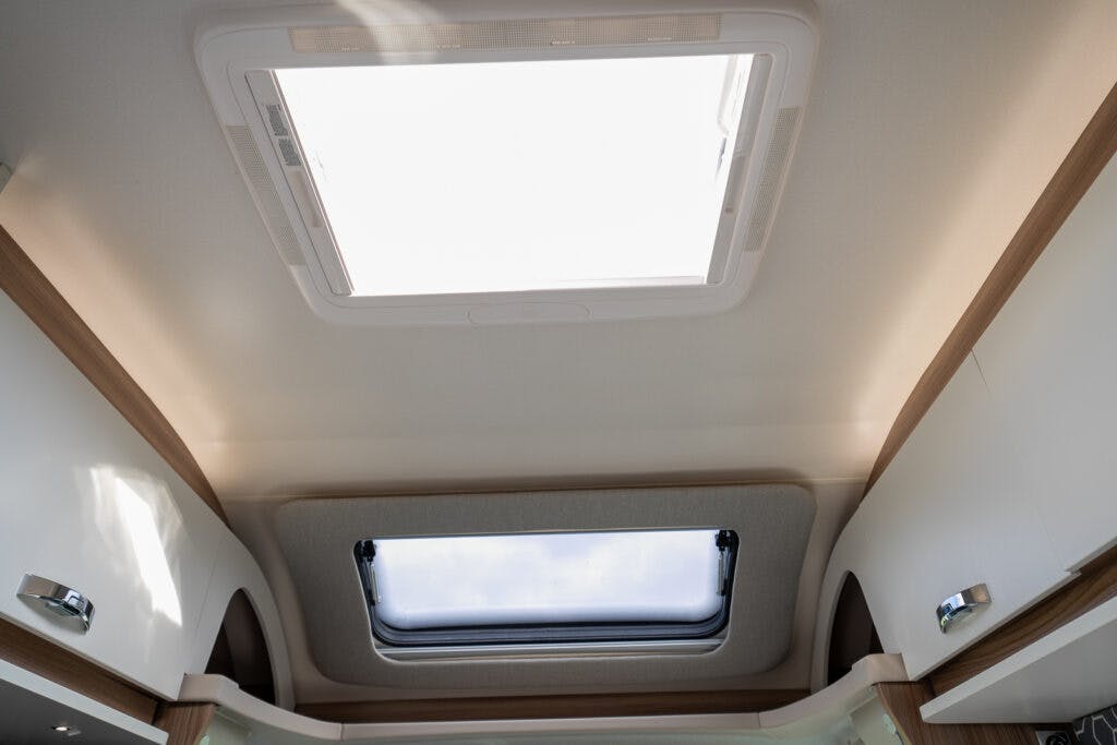A view of the ceiling inside a 2017 Swift Escape 664 RV, showing two skylights. The upper skylight is larger and fully closed, while the lower one is partially open. Surrounding the skylights are white and wooden interior panels.