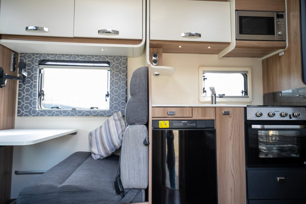 Interior of the 2017 Swift Escape 664 features a small dining area with a table and cushioned bench seating, kitchen appliances including a microwave, sink, dishwasher, and oven. The space is efficiently organized with light wood and white cabinetry.