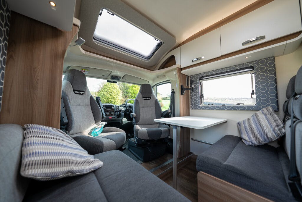 Interior view of a modern 2017 Swift Escape 664 motorhome showcasing a seating area with grey cushions and throw pillows, a table, and a steering compartment at the front. The space is lit by natural light coming through large windows and a skylight.