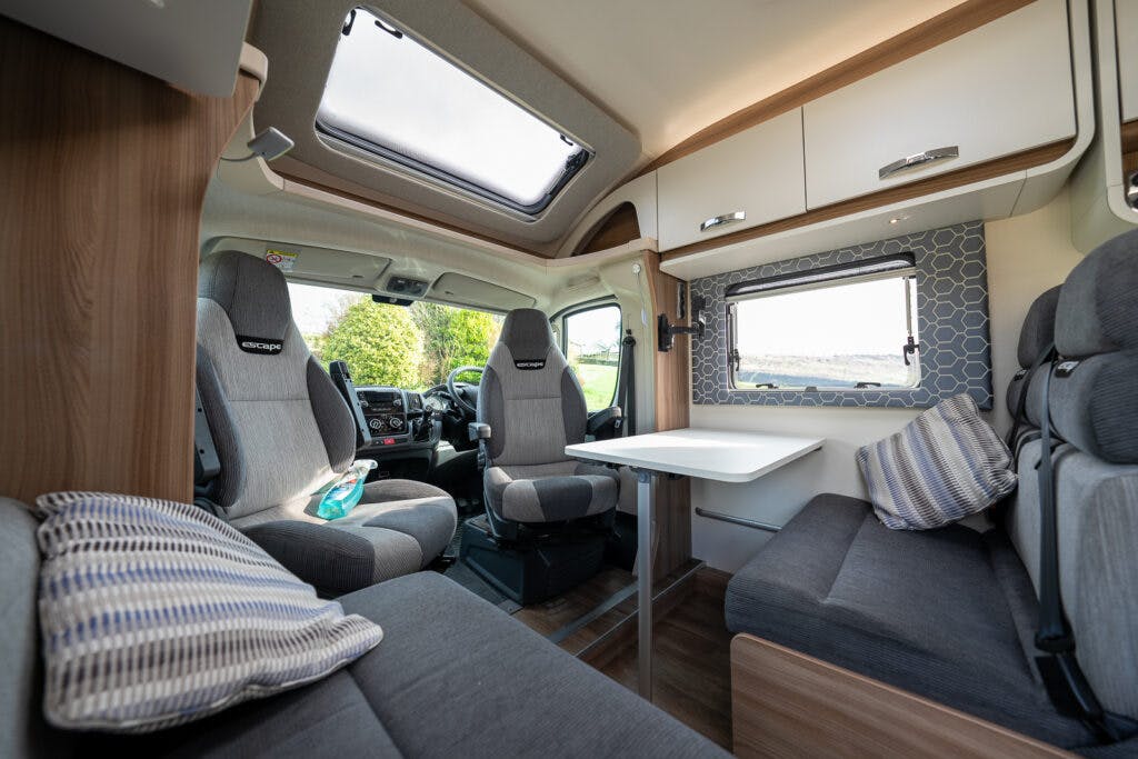 Interior of a 2017 Swift Escape 664 showing a seating area with two gray captain’s chairs and a gray bench with a striped cushion. There is a white fold-out table in the center and a large window with a blue geometric patterned shade. A skylight is overhead.