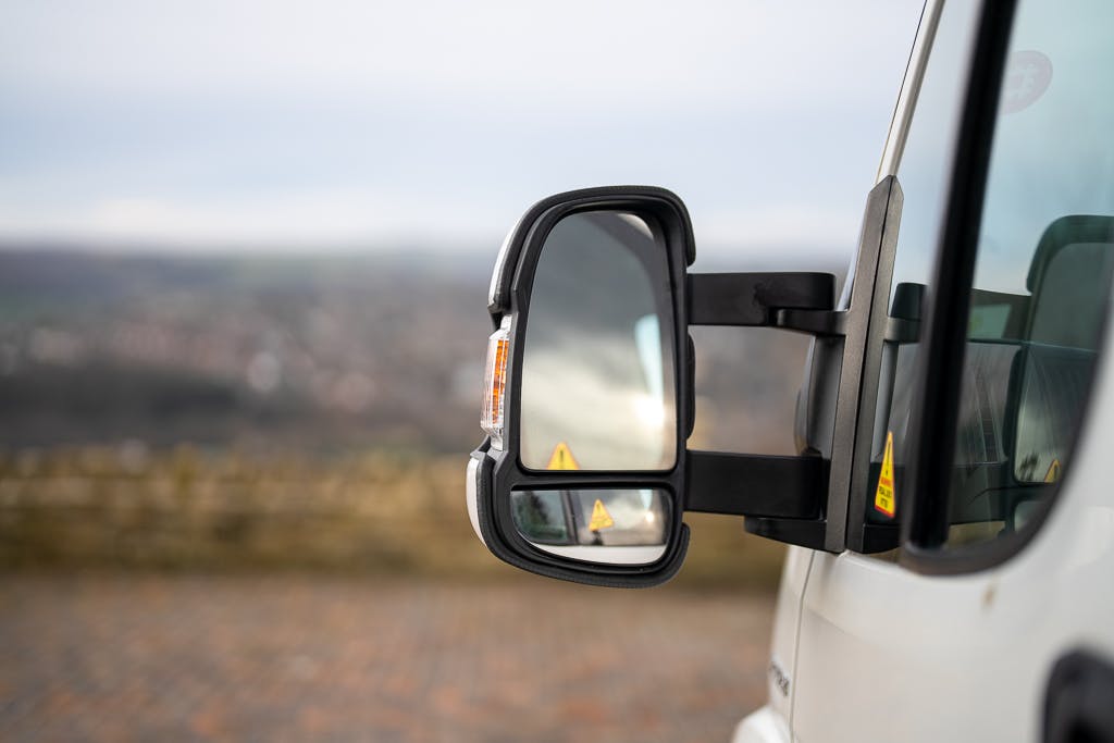 Close-up of the 2007 Auto-Sleepers Sigma EL's side mirror, showcasing its multiple reflective surfaces. The white vehicle features a black-framed mirror. In the blurred background, distant hills and sky hint at an outdoor setting.
