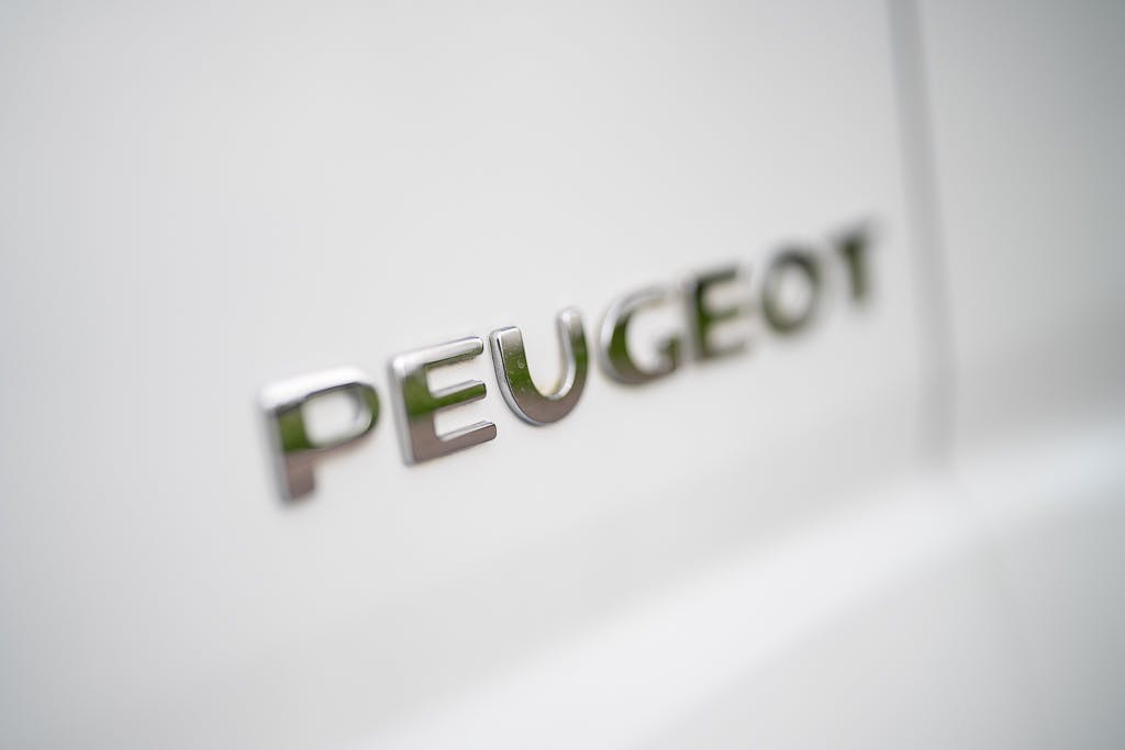 Close-up of the silver "Peugeot" logo on a white 2007 Auto-Sleepers Sigma EL. The polished metal letters stand out against the vehicle's exterior, with the white surface providing a contrasting background for the shiny emblem.