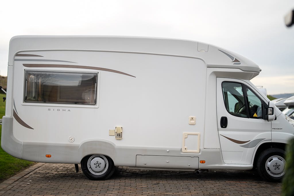 A white 2007 Auto-Sleepers Sigma EL motorhome with the word "SIGMA" on its side is parked on a paved surface. The vehicle features a side window, a door, and storage compartments. The background shows a cloudy sky and parts of other vehicles.