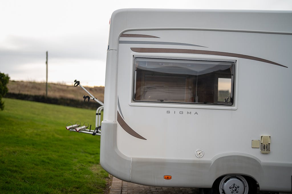 A white 2007 Auto-Sleepers Sigma EL is parked on a grassy field. The RV has a large side window, a small utility panel, and a bike rack attached to the rear. The sky is cloudy, and there is a wooden fence in the background.