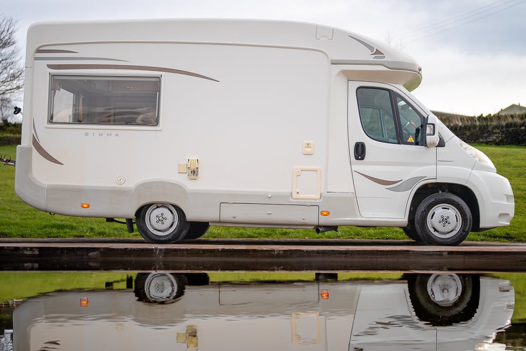 A 2007 Auto-Sleepers Sigma EL camper van with beige accents is parked on a concrete surface beside a small body of water reflecting the van. The camper has a window on its side and various external compartments. The background includes green grass and a cloudy sky.