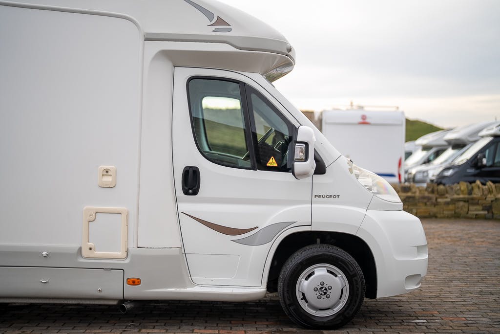 Side view of a white 2007 Auto-Sleepers Sigma EL motorhome parked on a paved surface. The motorhome has a Peugeot logo near the driver's door and features a small window and storage compartment on the side. Other motorhomes are visible in the background under a cloudy sky.