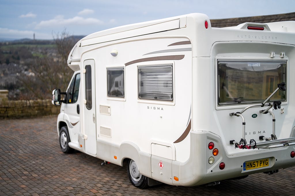 A 2007 Auto-Sleepers Sigma EL motorhome is parked on a paved area with a picturesque countryside backdrop. The white vehicle features a rear window, a bike rack, and an external storage compartment. Its license plate reads "HN57 PYG.