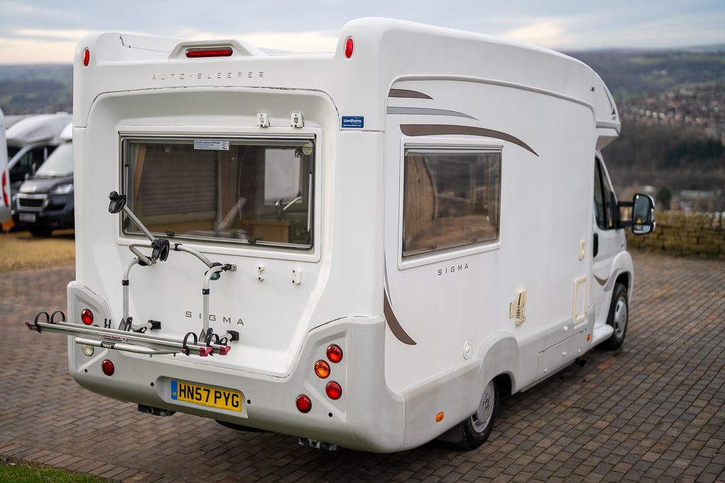 A white 2007 Auto-Sleepers Sigma EL camper van is parked on a paved surface. The back of the camper features a bicycle rack, windows, and taillights. The license plate reads "HN57 PVG." The background includes other vehicles and a scenic view.