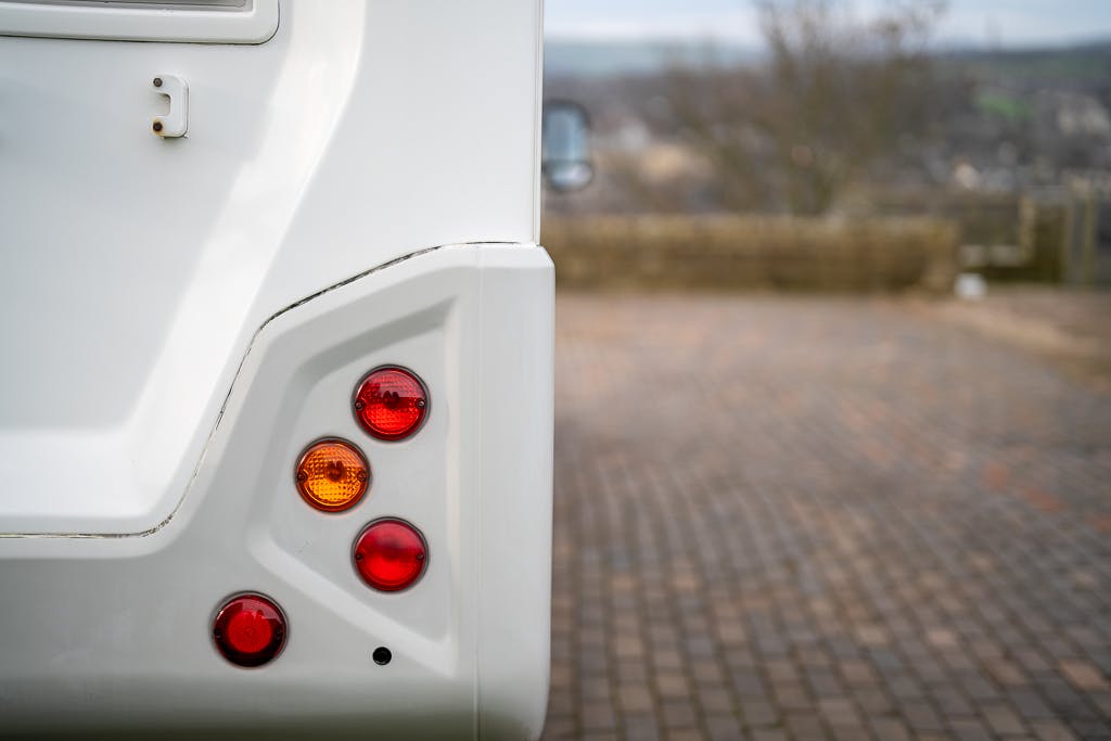 Close-up view of the back corner of a white 2007 Auto-Sleepers Sigma EL, focusing on its tail lights, which include red and amber colored lenses. The vehicle is parked on a cobblestone surface with a blurred background of a rural landscape.