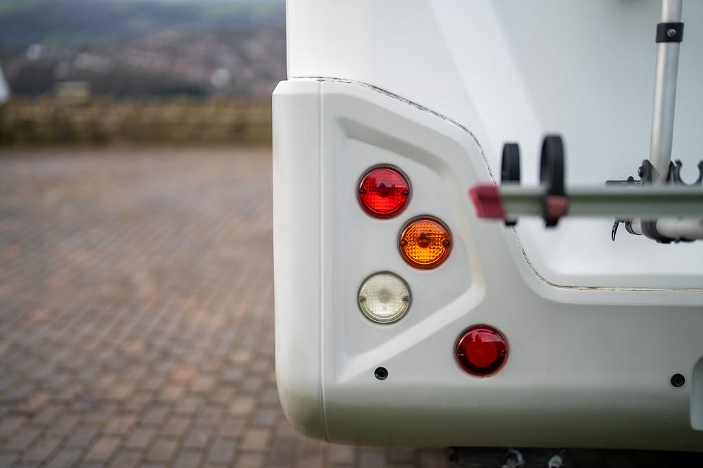 Close-up of the rear corner of a white 2007 Auto-Sleepers Sigma EL, showcasing its taillights. The vehicle is parked on a brick-paved surface with a blurred background, potentially indicating a scenic location. A part of a bike rack is visible attached to the vehicle.