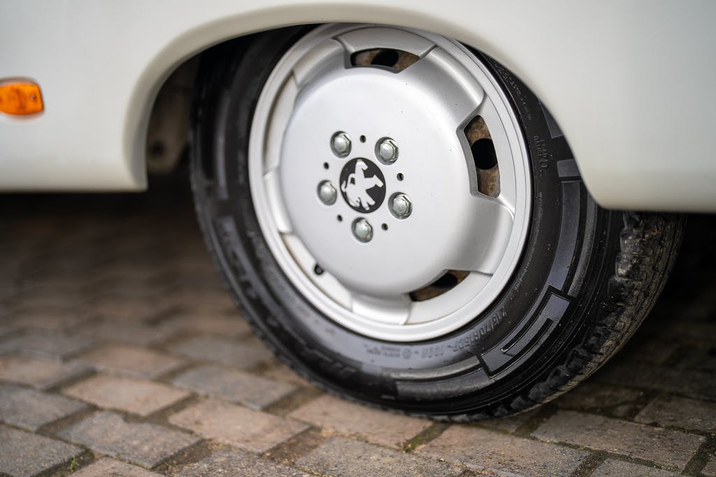 Close-up of a car tire and wheel on a stone-paved ground. The black tire shows clear tread, while the silver wheel features a lion logo in the center. A glimpse of the 2007 Auto-Sleepers Sigma EL's body and an orange side marker light are also slightly visible.