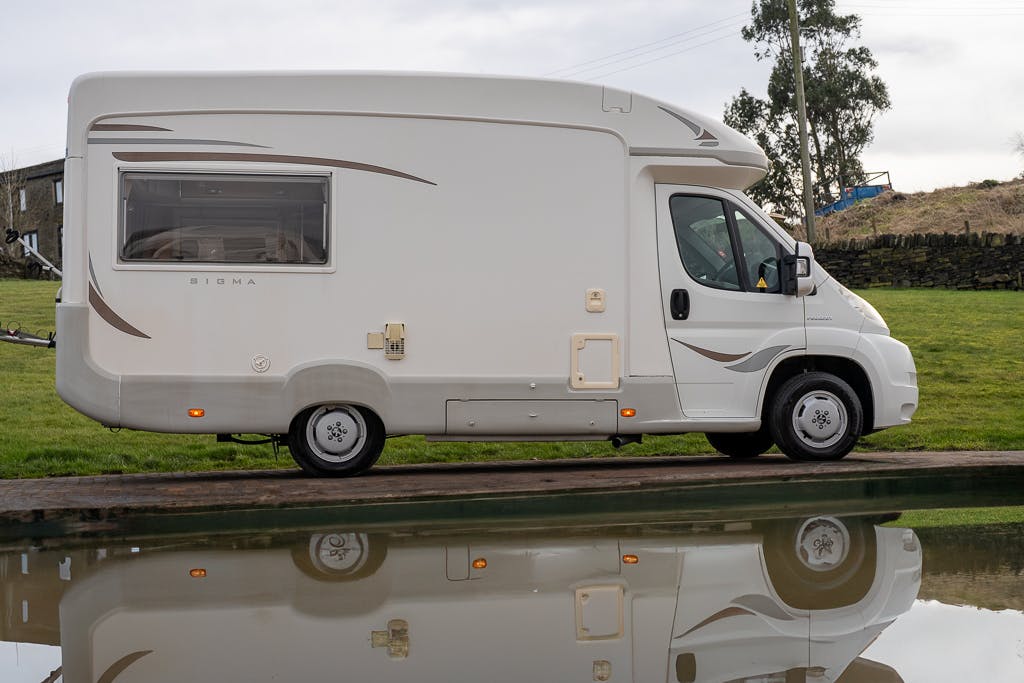 A white motorhome, identifiable as a 2007 Auto-Sleepers Sigma EL, is parked beside a calm, reflective body of water on a grassy area. The vehicle has beige decorative stripes along its side, and electrical hookups and windows are visible on the RV.
