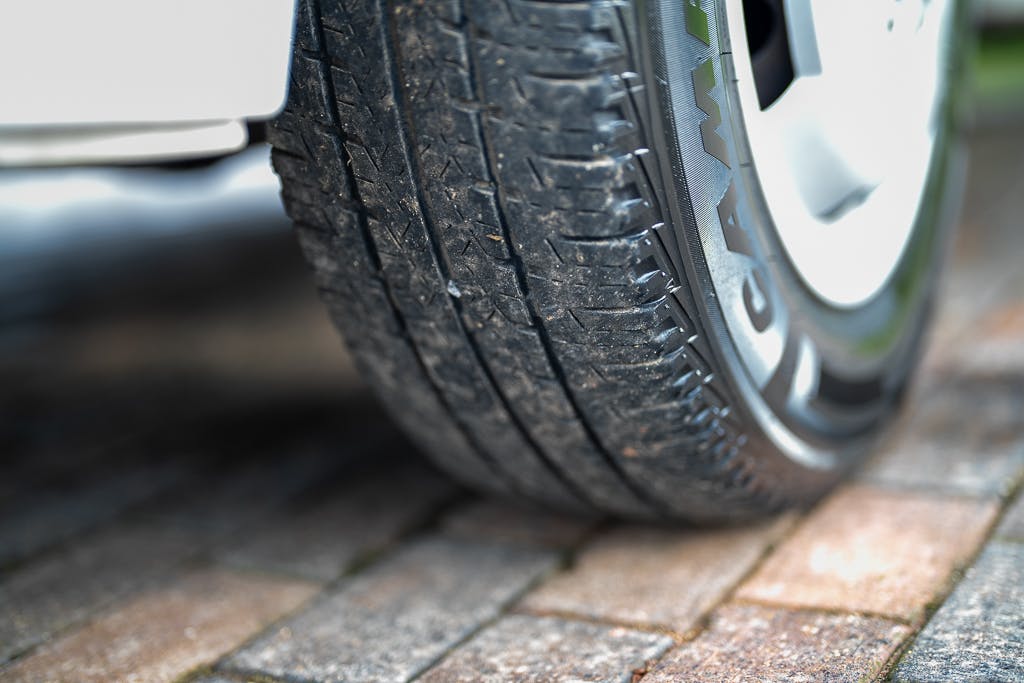 Close-up view of a car tire with visible tread patterns, positioned on a brick-paved surface next to the 2007 Auto-Sleepers Sigma EL. The tire appears to be in good condition with some dirt and debris lodged in the treads. Part of the vehicle’s white wheel well is also visible in the background.