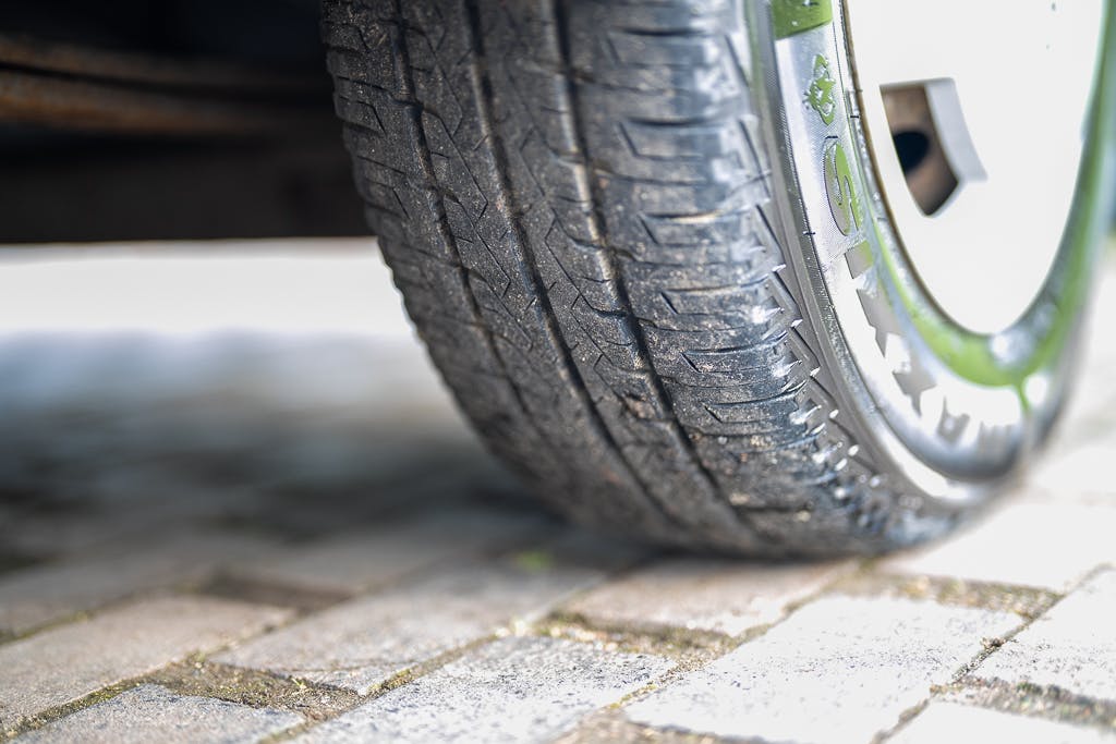 Close-up of a 2007 Auto-Sleepers Sigma EL's tire making contact with a paved surface. The tire shows signs of wear and has treads designed for traction. The wheel is partially visible, and the ground is composed of interlocking stone pavers.