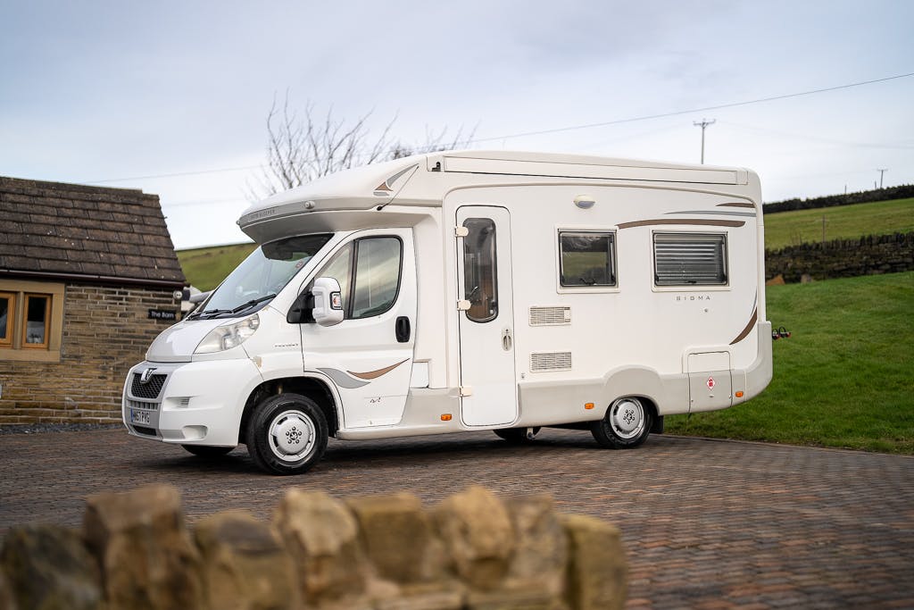 A 2007 Auto-Sleepers Sigma EL motorhome is parked on a paved surface near a small building. It has a cab-over section and side windows. The surrounding area consists of a field with short grass and a stone wall in the foreground. The sky is overcast.
