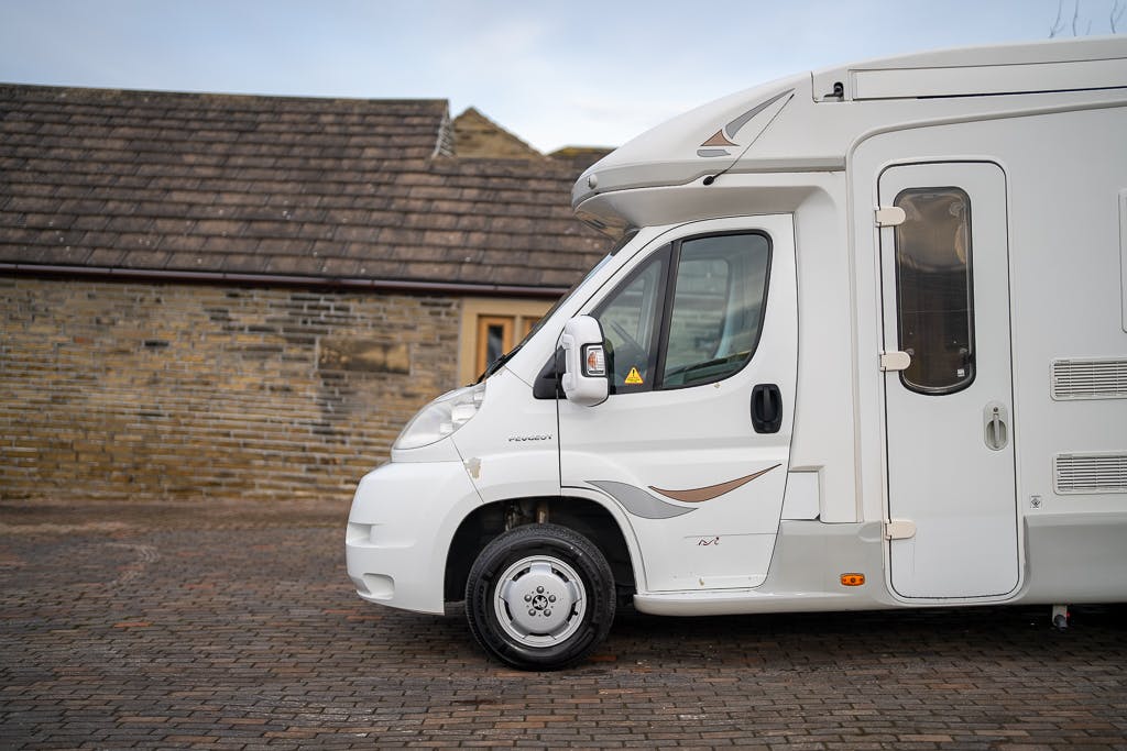 A 2007 Auto-Sleepers Sigma EL motorhome is parked on a cobblestone driveway in front of a stone building with a steep, shingled roof. The focus is on the side profile of the motorhome, showing the door and side mirror. The sky is partly cloudy.