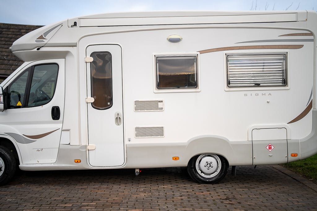 A white 2007 Auto-Sleepers Sigma EL camper van is parked on a paved surface. It features the "Sigma" brand name on its side, a side entrance door with a window, several ventilation panels, and a side window with shutters. Additionally, the vehicle is equipped with a small ladder on its rear side.