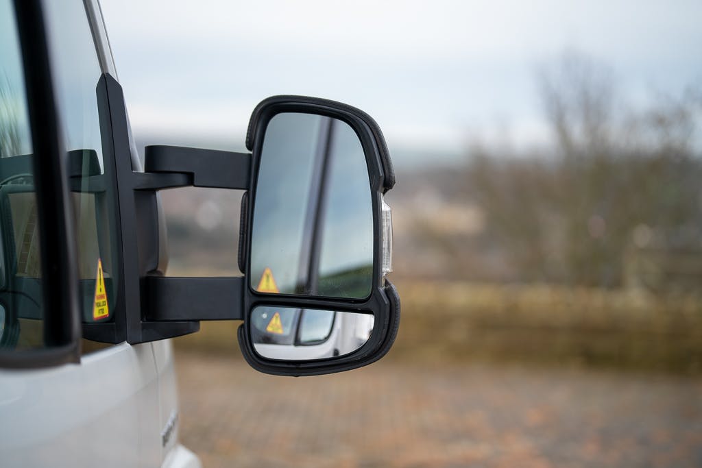A close-up view of a large vehicle's side mirror. The side mirror reflects portions of the 2007 Auto-Sleepers Sigma EL and a blurry background of trees and sky. Yellow warning labels are visible on the mirror. The image focuses on the mirror, with the rest of the vehicle partially visible.