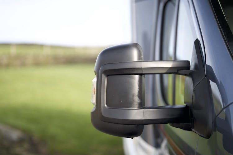 Close-up of a black side mirror on a 2014 Auto-Trail Imala 715 Lowline with a blurred green landscape in the background. The mirror's design is detailed, showing two support arms and a turn signal indicator. The image focuses on the mirror, with a minimal portion of the vehicle visible.