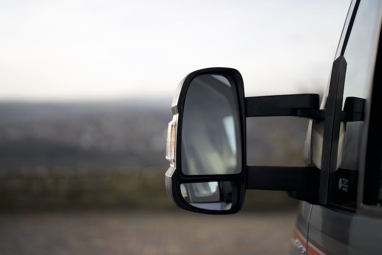 A close-up view of a 2014 Auto-Trail Imala 715 Lowline's side mirror, showing its reflective surface. The background is blurred, with an indistinct landscape and a cloudy sky visible. The vehicle appears to be parked on a stone-paved area.