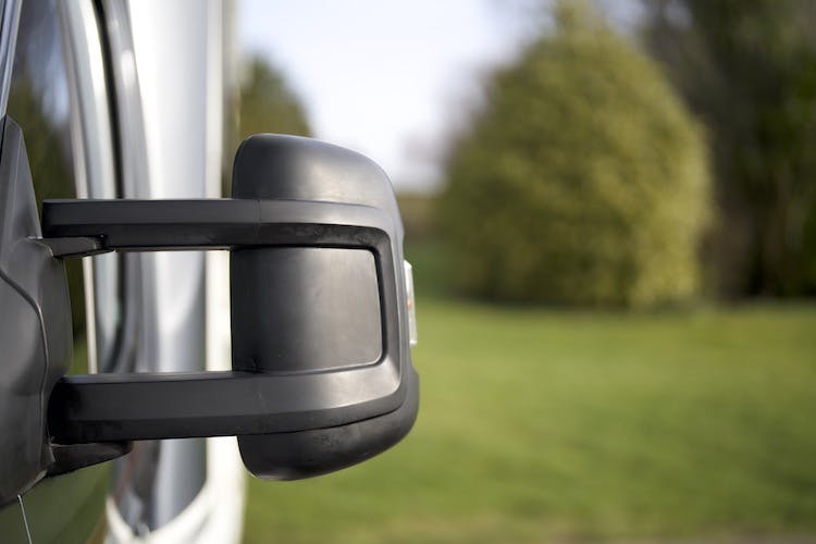 Close-up of a black side mirror attached to the exterior of a white 2014 Auto-Trail Imala 715 Lowline, with a blurred green lawn and trees in the background. The mirror is rectangular and positioned perpendicular to the side of the vehicle.