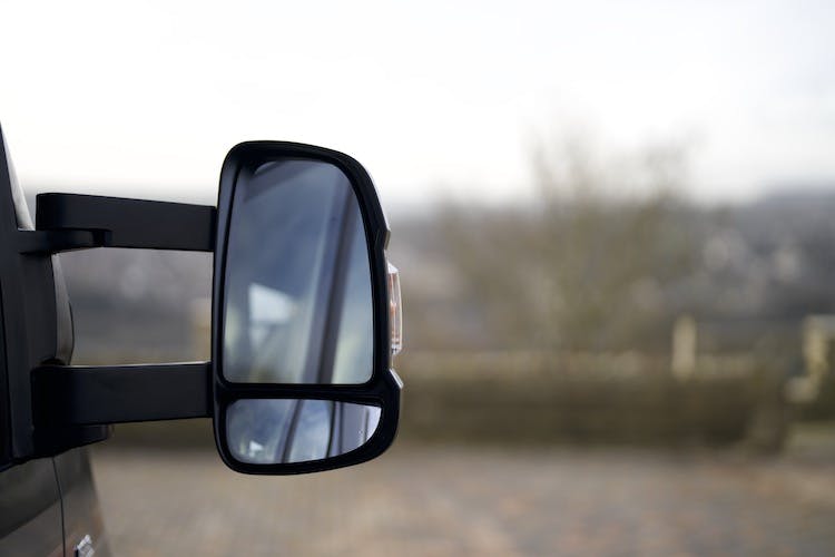 Close-up of the side mirror of a black 2014 Auto-Trail Imala 715 Lowline, with an out-of-focus background showing a landscape and trees. The mirror reflects part of the vehicle and a hint of the surroundings.