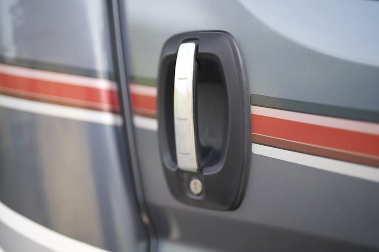 Close-up of a silver door handle on a gray 2014 Auto-Trail Imala 715 Lowline. The door features horizontal stripes in red, white, and black running across it. The keyhole is visible below the handle.
