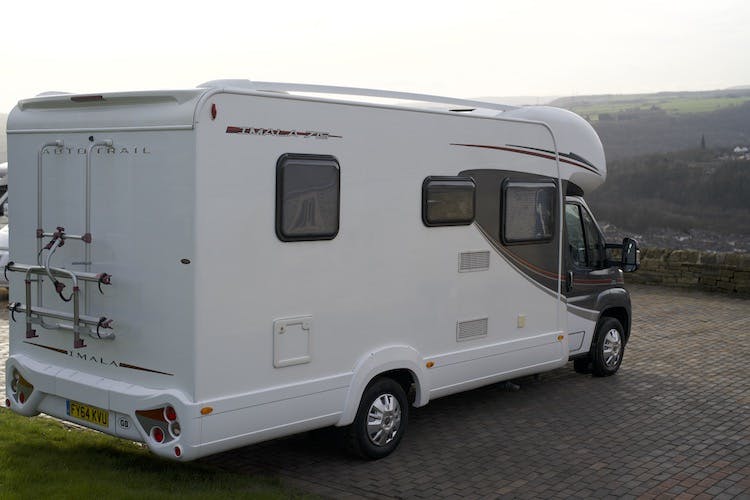 A 2014 Auto-Trail Imala 715 Lowline motorhome is parked on a paved driveway overlooking a scenic countryside. The back of the motorhome has a bike rack, and the side features a window and storage compartments. The background shows a panoramic view of rolling hills and fields.