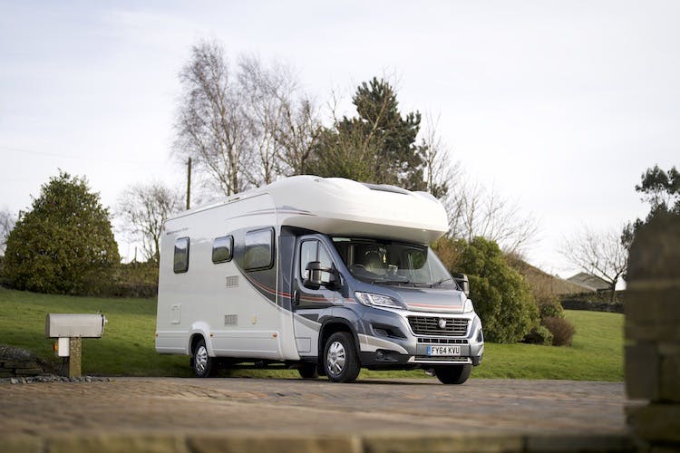 The image shows a 2014 Auto-Trail Imala 715 Lowline motorhome parked on a paved area in front of a grassy landscape with several trees and bushes in the background. The white motorhome, accented with darker tones, bears the license plate number FY64 KLU.