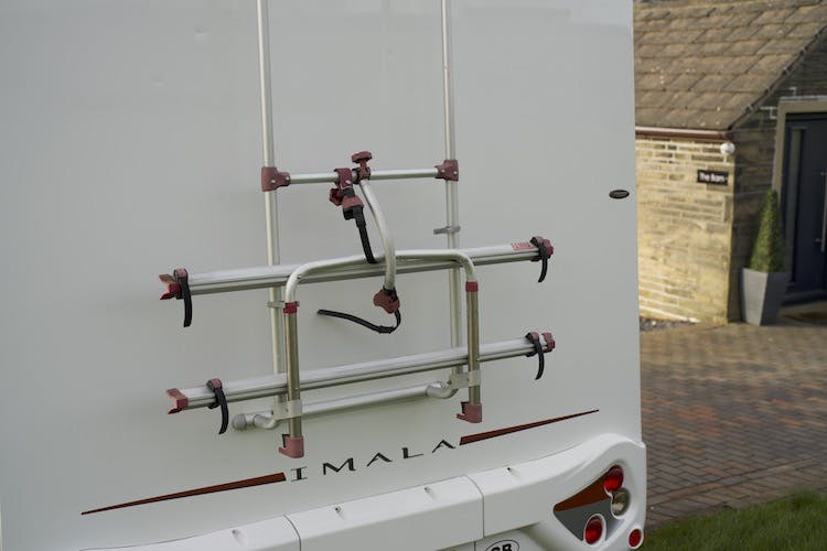 The image shows the rear side of a white 2014 Auto-Trail Imala 715 Lowline RV. Attached to the back is an empty bicycle rack. The brand "IMALA" is printed on the lower part of the vehicle's rear. A house is visible in the background.