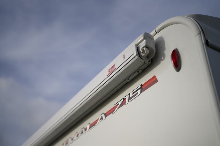 Close-up of the upper rear corner of a white 2014 Auto-Trail Imala 715 Lowline camper vehicle with a small red light. The Fiamma F45s awning is mounted near the top, and the text "TMCALA-715" is visible below it. The sky in the background is partly cloudy.