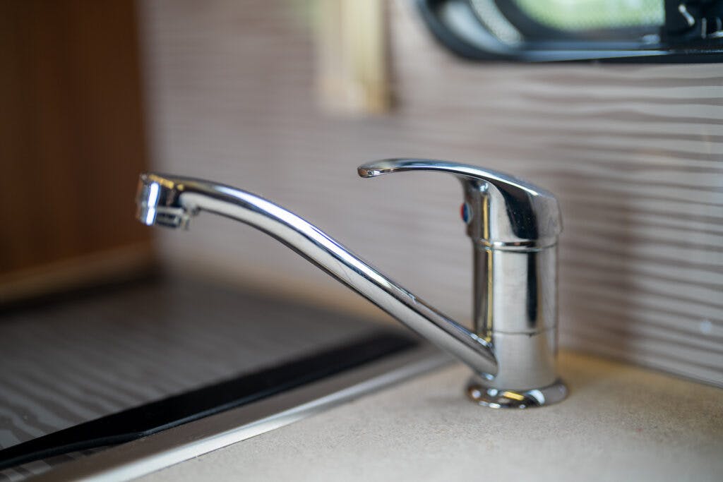 A close-up of a modern, silver-colored kitchen faucet with a single lever handle on a beige countertop. The background shows part of a stainless steel sink and a tiled backsplash in a stylish kitchen setting reminiscent of the sleek design of the 2014 Auto-Trail Imala 715 Lowline.
