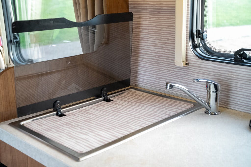 The image shows a kitchen sink in a 2014 Auto-Trail Imala 715 Lowline RV, with a protective cover over the sink basin. A modern faucet is positioned to the right of the covered sink. The interior is decorated with neutral tones, and the window next to the sink provides an outward view.