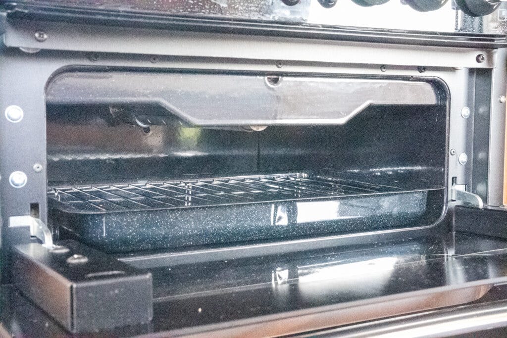 An open oven door in the 2014 Auto-Trail Imala 715 Lowline reveals the interior, featuring a wire rack positioned in the middle and a black, rectangular baking pan on the rack. The oven appears clean and unused.