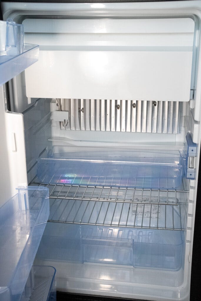 An open, empty refrigerator with a compact design and white interior. It contains several clear blue plastic shelves and drawers. The back wall showcases a silver panel. This modern fridge would be right at home in a 2014 Auto-Trail Imala 715 Lowline motorhome, adding convenience to any journey.