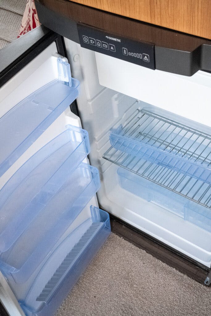 An open refrigerator inside the 2014 Auto-Trail Imala 715 Lowline displays empty, transparent blue door shelves, a clear drawer at the bottom, and wire racks in the main compartment. The door is partially open, revealing control buttons on the upper section. The floor beneath is carpeted.