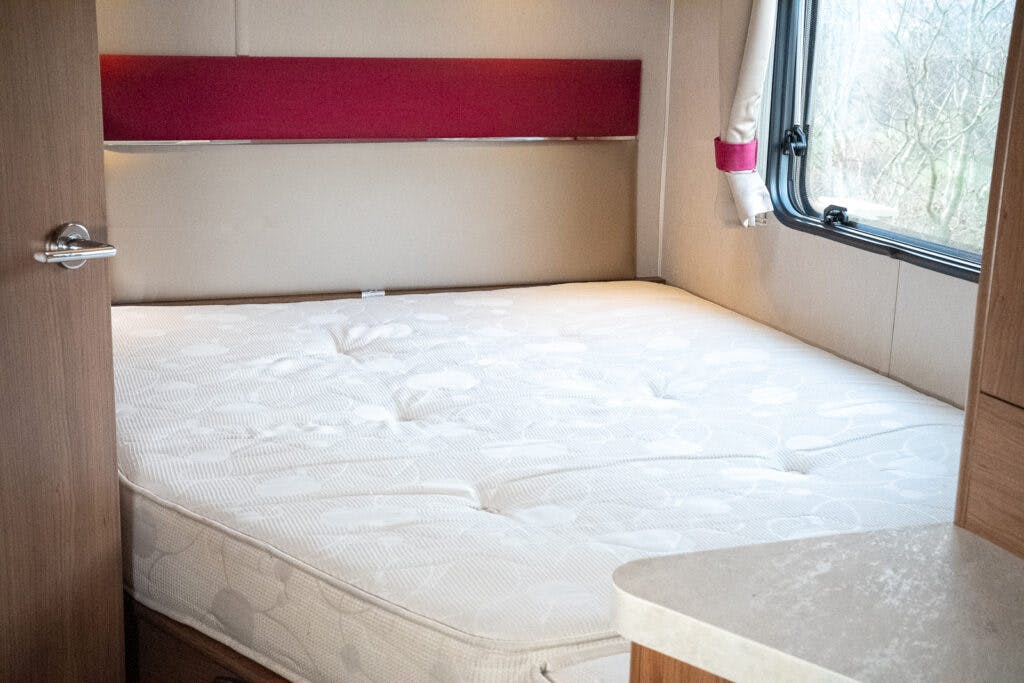 A small bedroom in a 2014 Auto-Trail Imala 715 Lowline RV features a neatly made bed with a white mattress. The room includes a large window with a curtain, a wooden door slightly ajar, and a red headboard panel. Light brown walls and a countertop are also visible.