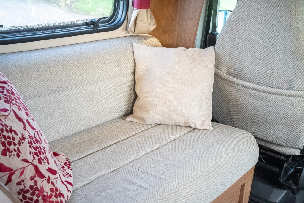 A beige cushion is placed on a light grey upholstered bench seat inside a 2014 Auto-Trail Imala 715 Lowline. Another cushion with a red floral pattern is partially visible to the left. The interior features neutral tones with a beige driver's seat in the background.