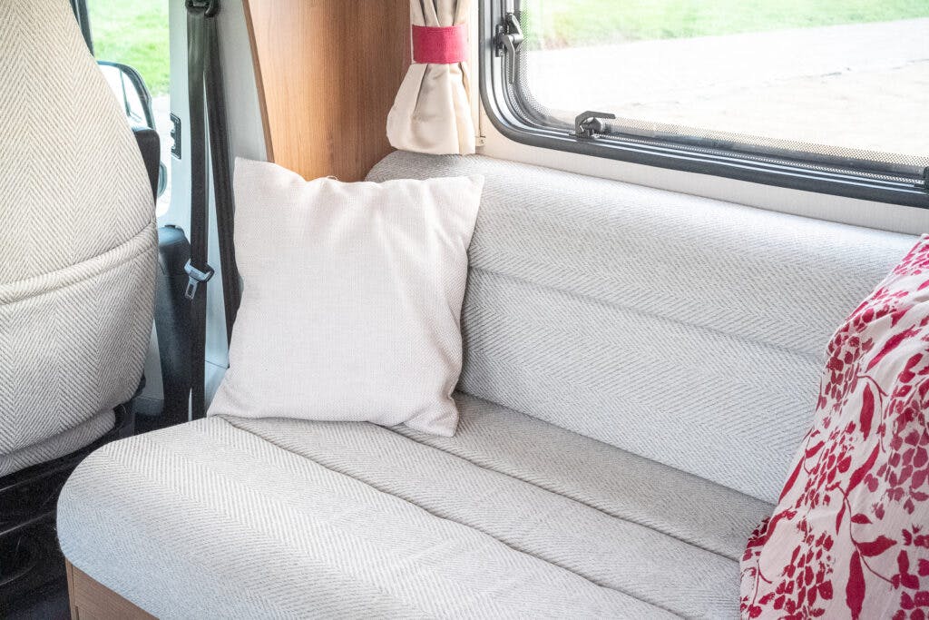 An interior view of the 2014 Auto-Trail Imala 715 Lowline camper van shows a light grey fabric sofa with a matching pillow. The window above the sofa has beige curtains tied back with red bands. The driver’s seat is partially visible on the left, and a red and white patterned fabric lies on the side.