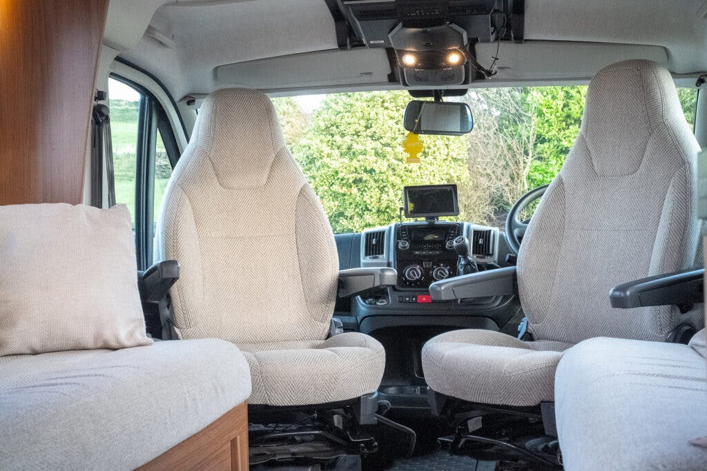 Interior of a 2014 Auto-Trail Imala 715 Lowline camper van showing two front seats, a central control dashboard with a mounted screen, and plush beige upholstery. The cozy, compact space is designed for traveling comfort, with natural light streaming in from the windows.