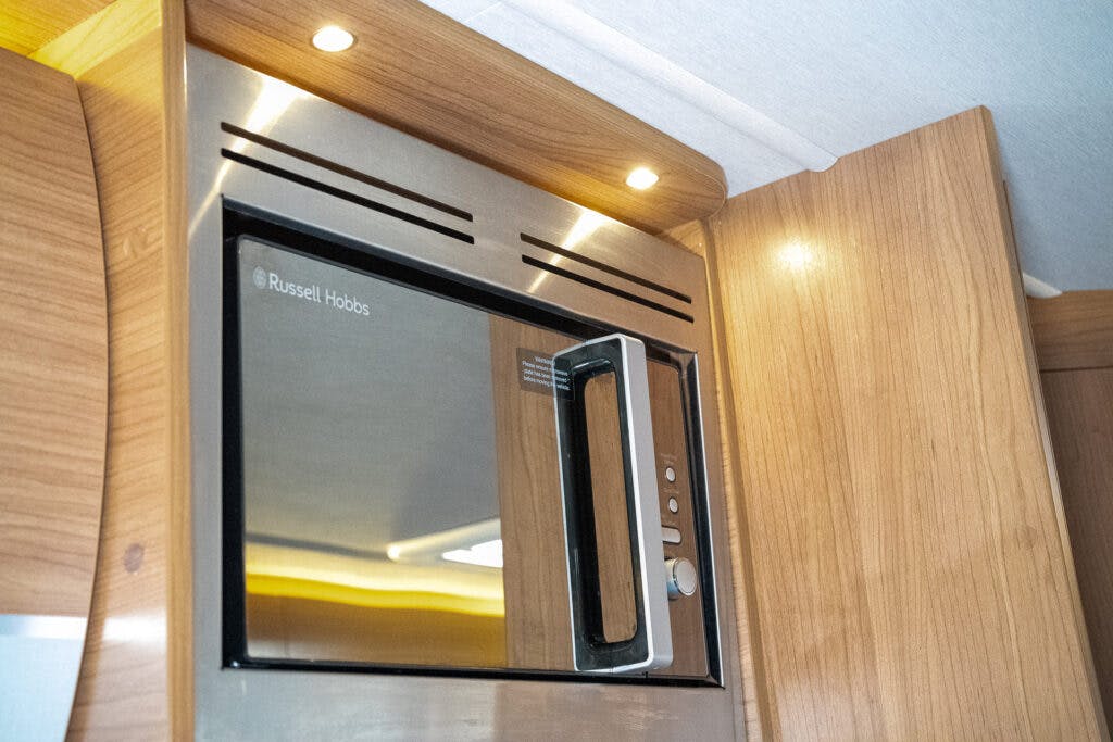 A stainless steel Russell Hobbs microwave is built into a wooden kitchen cabinet in the 2014 Auto-Trail Imala 715 Lowline. The microwave has a black glass door with a handle and buttons on the right side. The cabinet features recessed lighting above the microwave, adding a touch of elegance.