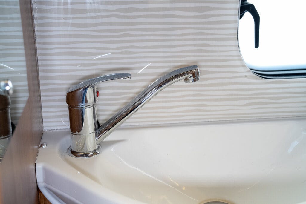 A chrome kitchen faucet extends over a white sink, reminiscent of the sleek design found in a 2014 Auto-Trail Imala 715 Lowline. The background features a white and gray striped pattern, with a partial view of a window on the right side.