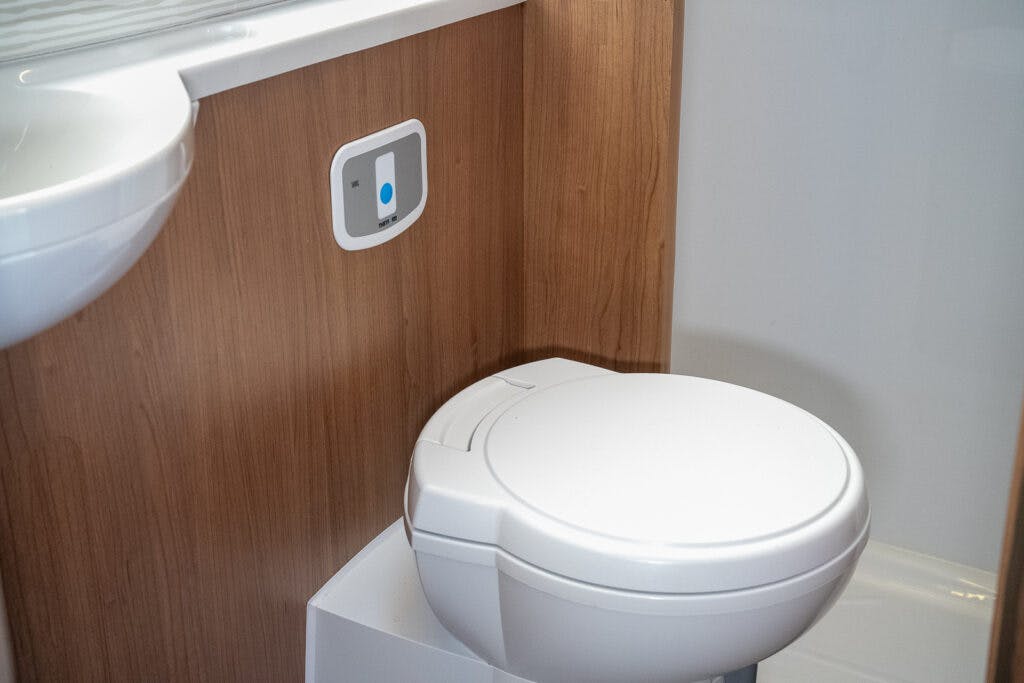 A white toilet with a round seat is installed against a wooden panel wall, resembling the interior of a 2014 Auto-Trail Imala 715 Lowline. A small control panel with a button is mounted on the wall next to the toilet. Part of a white sink is visible on the left side of the image.
