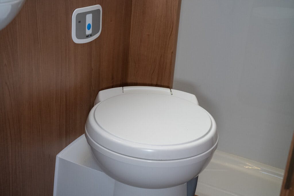 A white toilet with a closed lid is situated against a wall with wooden paneling inside the 2014 Auto-Trail Imala 715 Lowline. A rectangular control panel with a blue button is mounted on the wall next to the toilet. The floor and surrounding surfaces are clean, reflecting the compact efficiency of this motorhome.