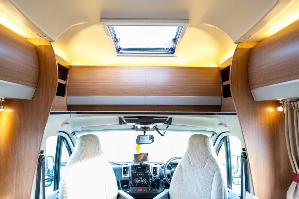 View from the rear of a 2014 Auto-Trail Imala 715 Lowline motorhome focusing on the front interior. The driver and passenger seats are visible, facing forward. The dashboard has a control panel and a hanging air freshener. Overhead storage compartments run above the seats, and a skylight is open.