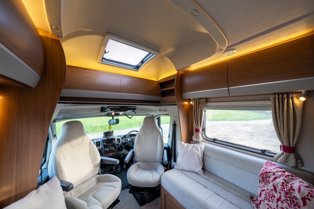 The interior of the 2014 Auto-Trail Imala 715 Lowline camper van is shown. It features two front seats, a skylight, and large windows with curtains. The seating area includes a compact couch with cushions. The space has wooden cabinetry and warm lighting.