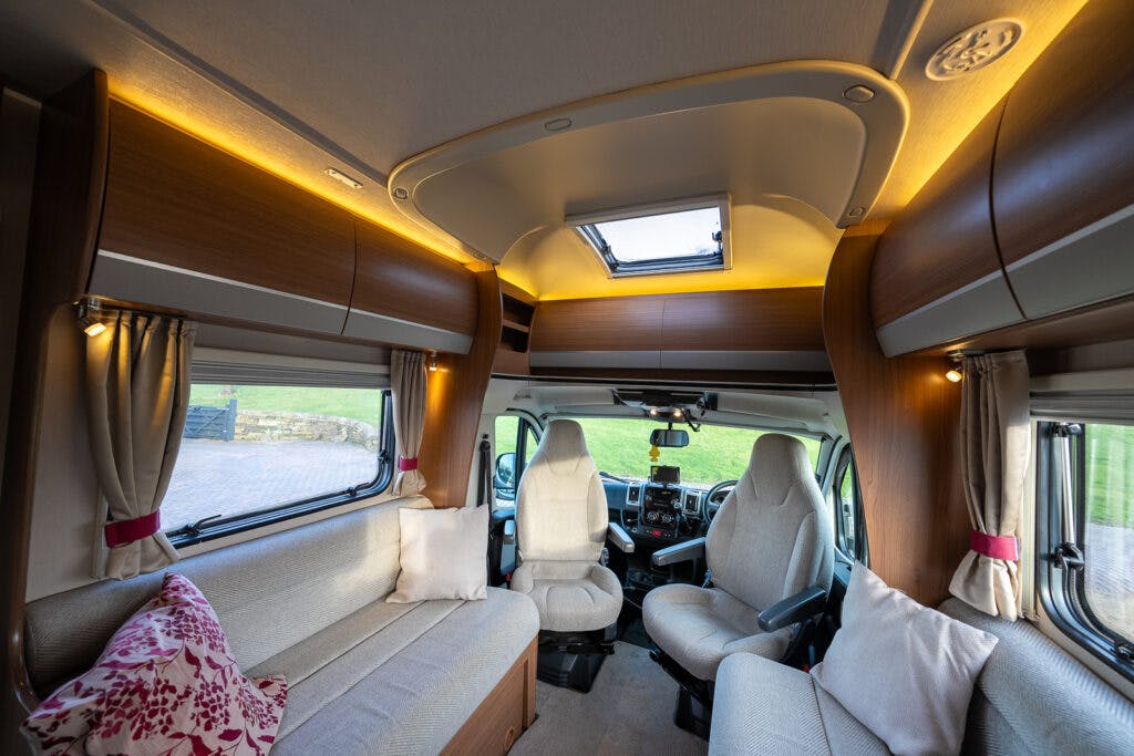 The interior of the 2014 Auto-Trail Imala 715 Lowline camper van features light brown wooden cabinets, beige seating, and a skylight. The space includes a driver and passenger seat in the front, alongside a small living area with overhead cabinets and a window with curtains.